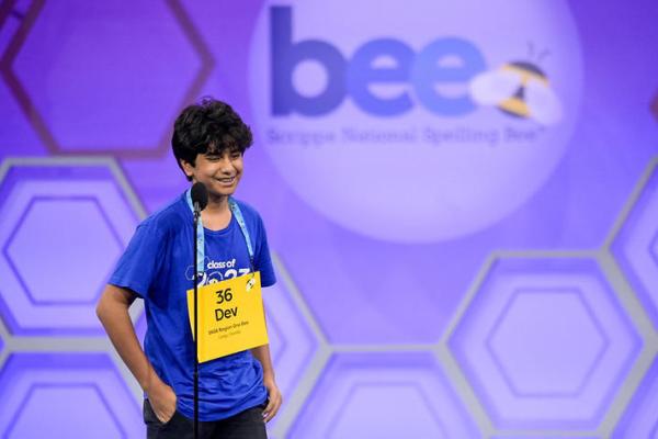 A boy standing on stage with a yellow placard hanging from his neck by a string. He is a participant in the Scripps National Spelling Bee. 