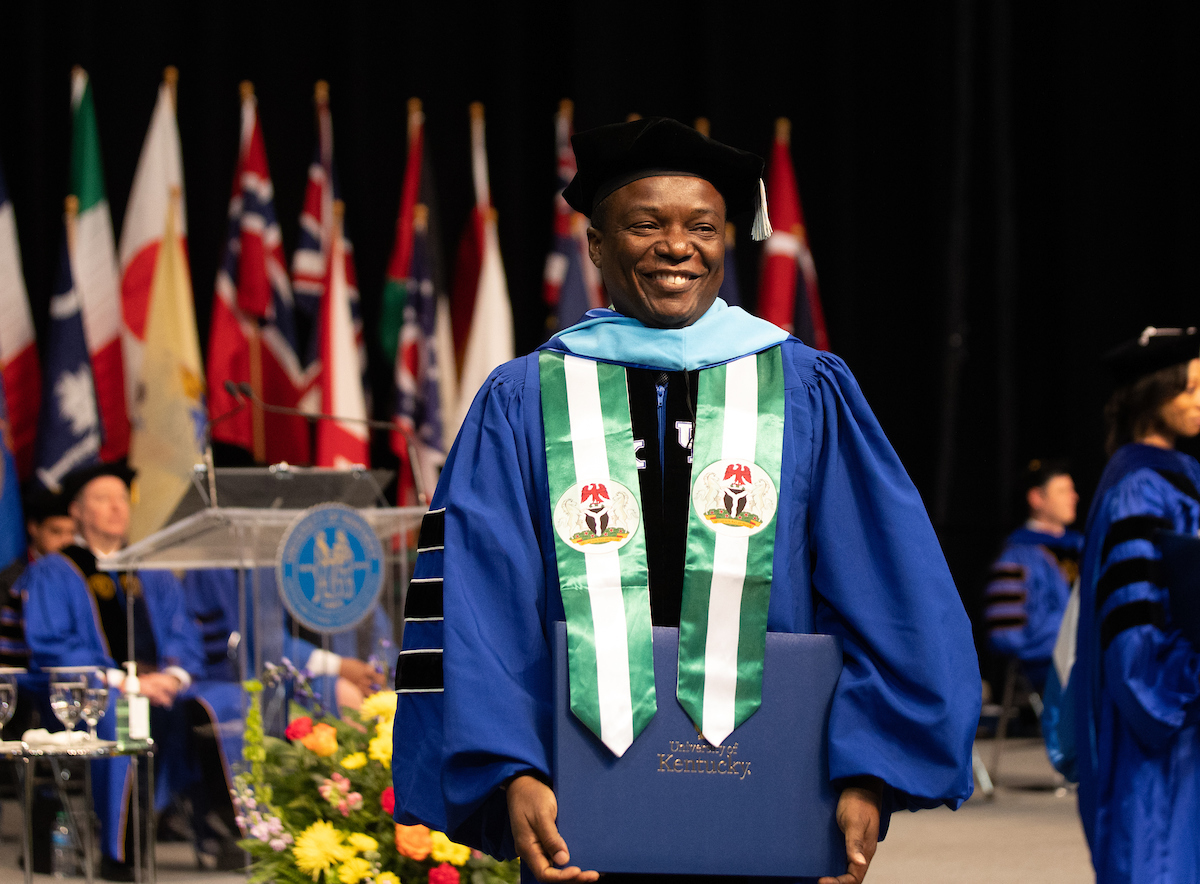 Photo of man wearing doctoral regalia. He is standing on stage at the University of Kentucky Commencement ceremony. There are flags on the stage behind him.