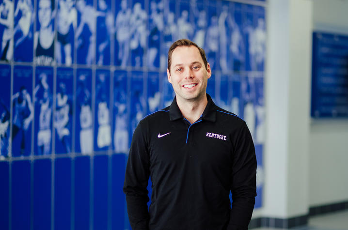 Man poses for a photo in front of a royal blue wall with images of student athletes on the wall