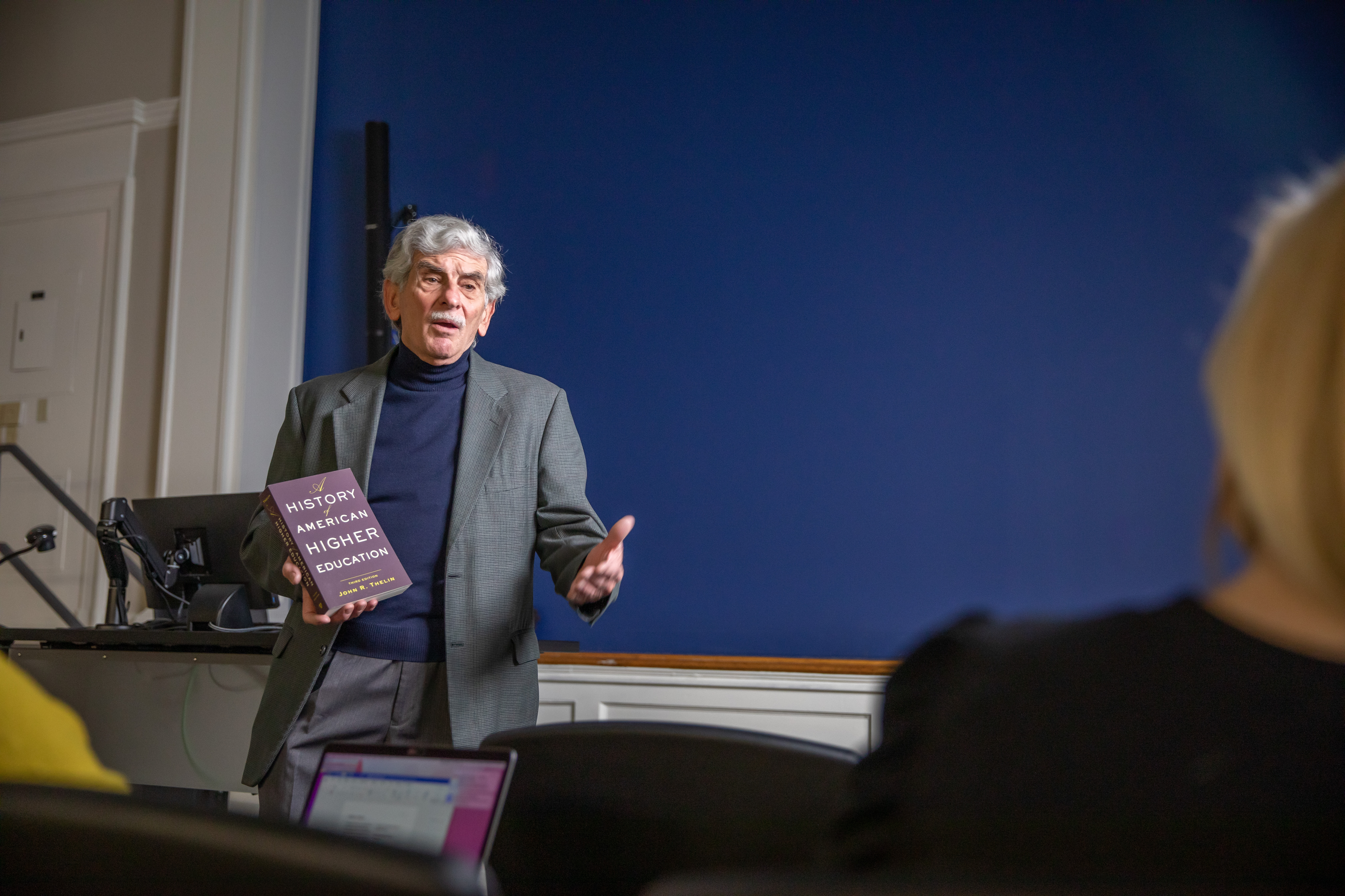 Photo of a man holding a textbook while speaking at the front of a room