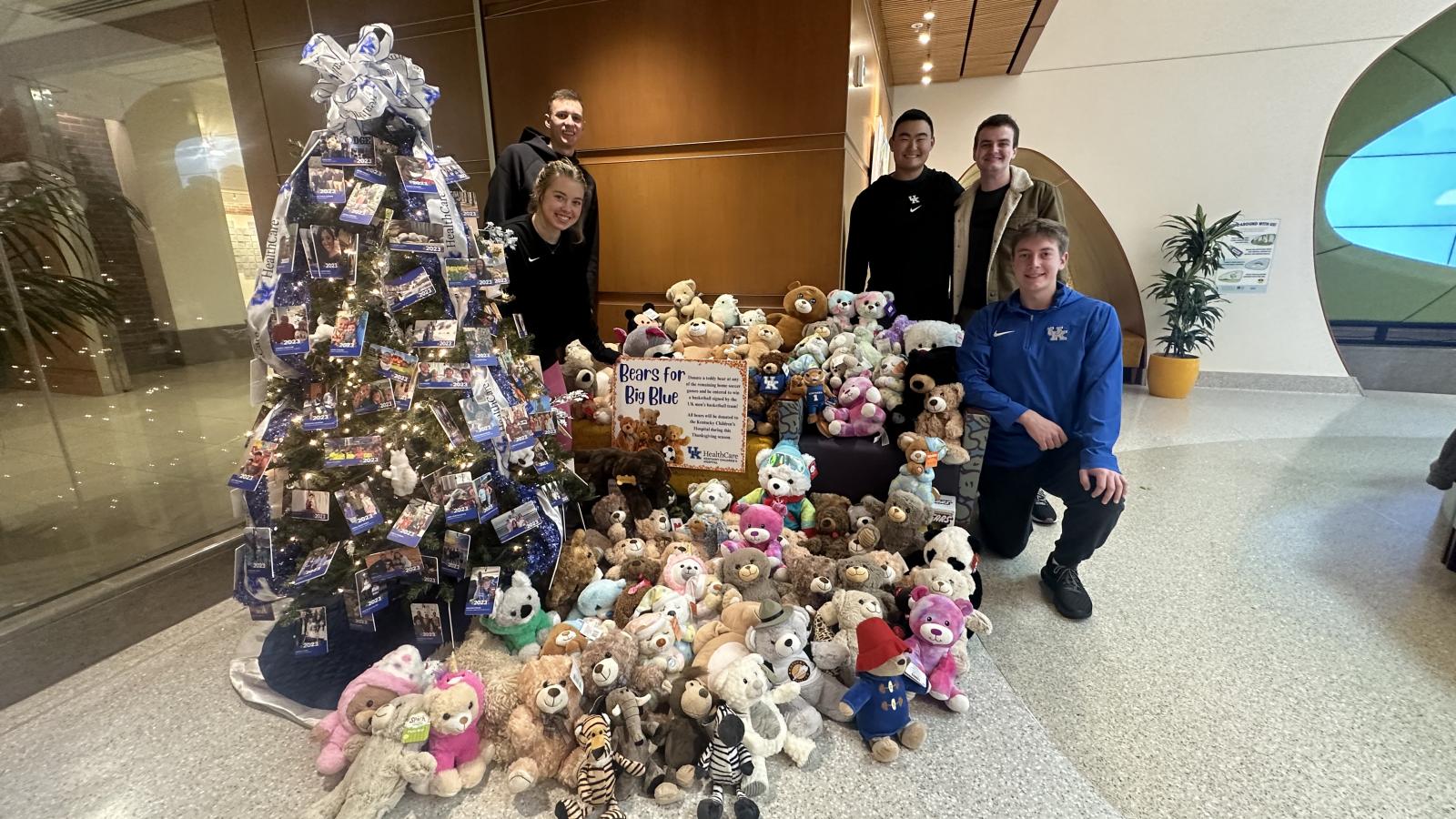 Five students standing next to a Christmas tree and stuffed animal donations for the children's hospital