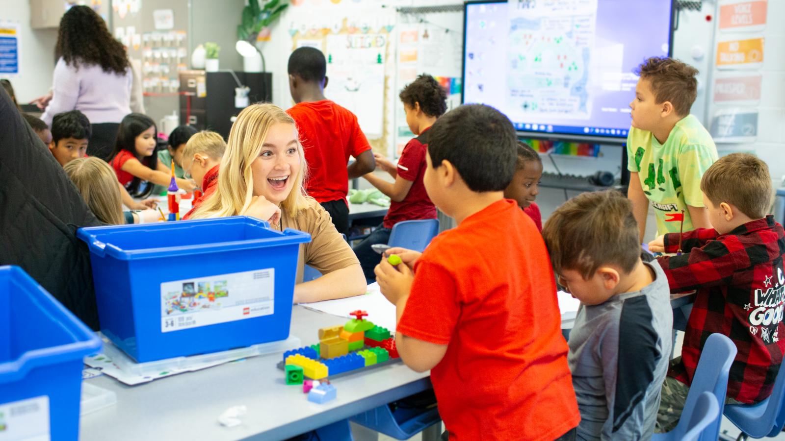 A UK student sits at a school desk with several children and interacts with them using Legos
