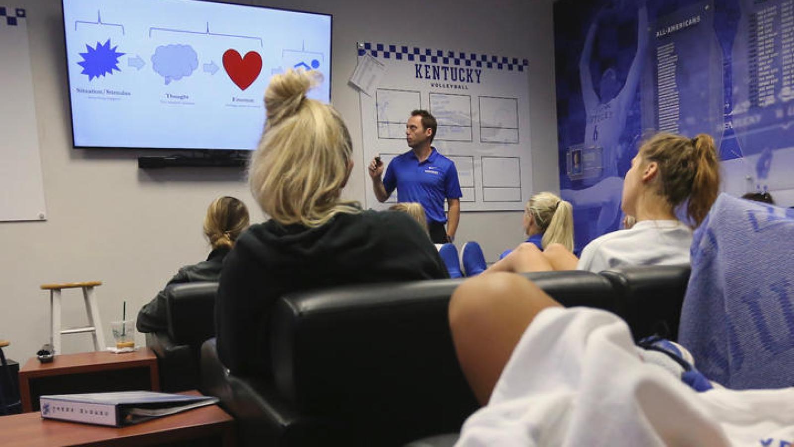 Man standing in front of room with presentation screen behind him. Student athletes are seated in chairs watching the presentation.