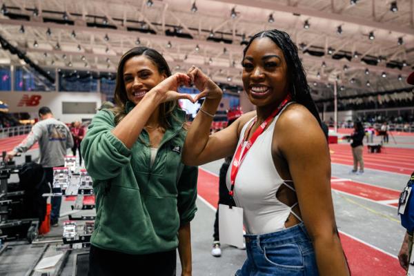Two women stand on the sidelines of an indoor track and field stadium. They are smiling and pressing their hands together to make the shape of a heart with their hands. 