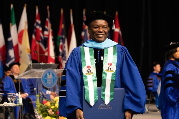 Photo of man wearing doctoral regalia. He is standing on stage at the University of Kentucky Commencement ceremony. There are flags on the stage behind him.