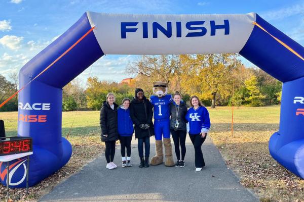 Five individuals plus the Kentucky Wildcat mascot stand at the 5k race finish line to pose for a photo