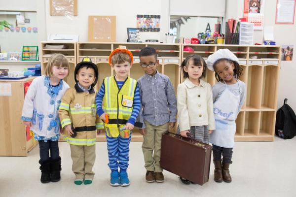 A group of six young boys and girls playing dress up in outfits such as a nurse, firefighter, doctor, construction worker, chef, and business professional.