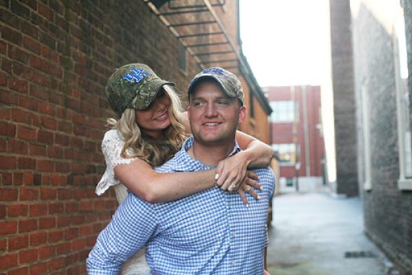 wife wrapping arms around husband, both wearing UK baseball caps