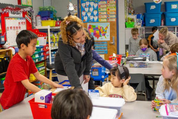 A UK student stands at a table in a second grade classroom and points to the students' work.