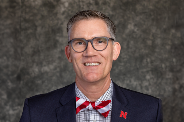 Photo of Nick Pace against grey background, wearing black jacket and red and white striped bow tie and glasses