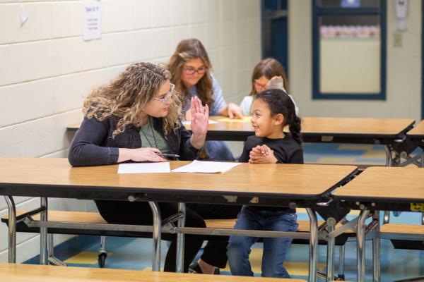 A UK student sits at a lunchroom table with a young student and gives her a high five.