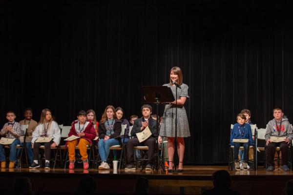 Photo of previous spelling bee. Students in the spelling bee are sitting in chairs on the stage behind a woman who is serving as emcee for the event, standing behind a podium and microphone.