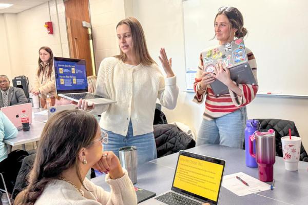 Two students stand next to a table holding laptop computers while making a presentation.