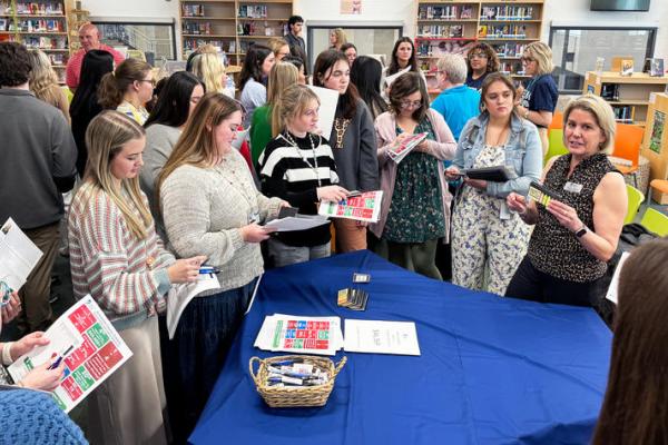 A group of students stands around a table with a blue table cloth. They are speaking to a hiring official from a nearby school district.