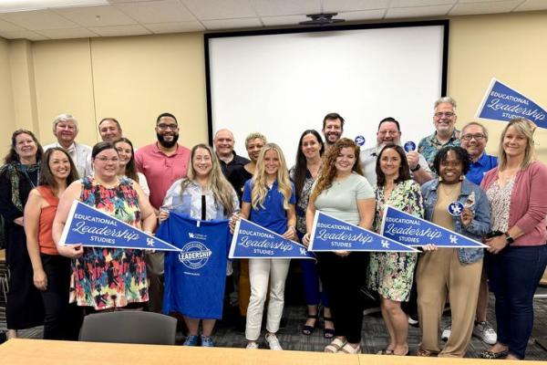 Large group of students and faculty stand in rows, holding up pennants and t-shirts that say UK College of Education