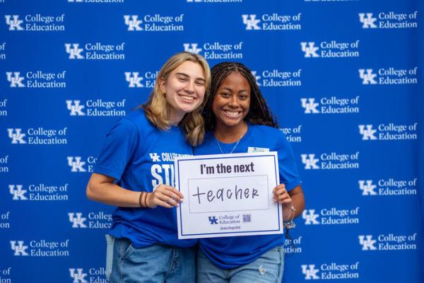Two women in blue tshirts stand holding a sign that says "I'm the next." They have filled in the blank with the word "teachers."