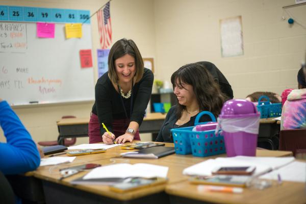 Teacher with brown hair wearing a black shirt is smiling and writing on a piece of paper that is on a desk in a classroom as a student with brown hair and dark blue shirt smiles and looks on
