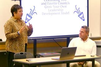 A woman is standing behind a small table. There is a PowerPoint presentation screen behind the woman. There is a man next to the woman. He is seated at the table and is listening to her speak.