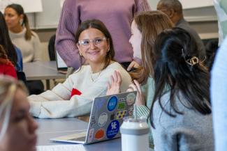 Group of students in a classroom, focus is on student in a white sweater wearing glasses and looking at a classmate smiling