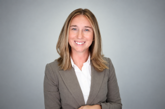 Woman in a brown blazer and white blouse smiling, standing in front of a grey background