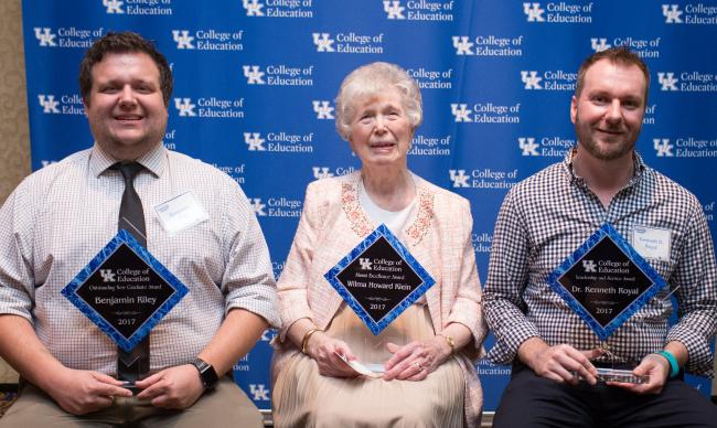 Three individuals sitting in a row. They are in front of a blue UK College of Education backdrop and are holding awards from a scholarship banquet.