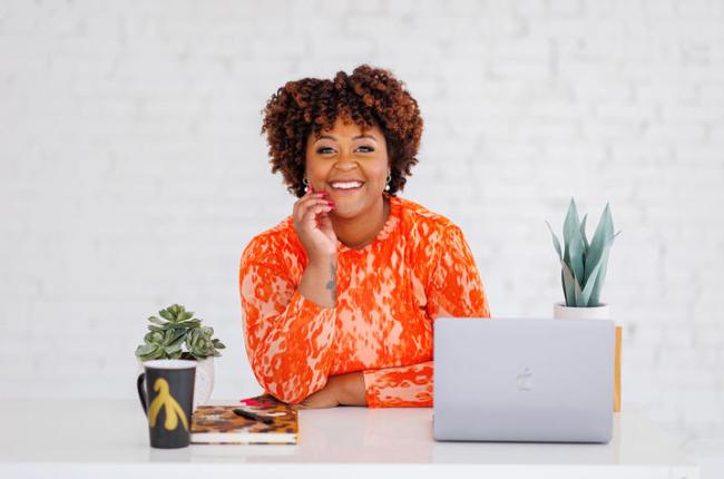 A woman posing for a professional photo. She is seated at a desk with her elbow on the desk and hand resting on her face. There is a laptop, coffee mug, a notebook, and plants on the desk.