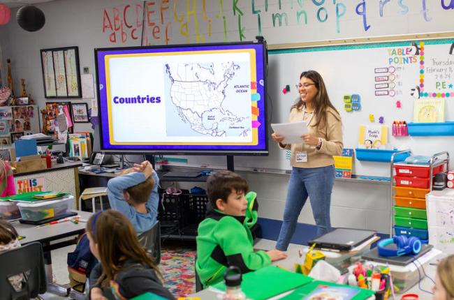 A college student stands at the front of an elementary classroom and is speaking with the students. There is a TV screen displaying a map of North America.