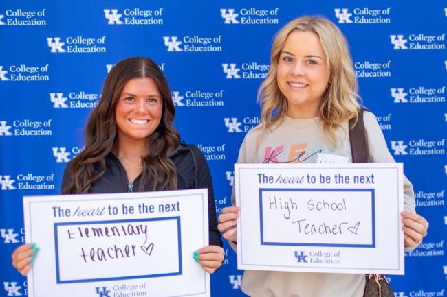 Two students standing in front of a UK step and repeat backdrop. One is holding a sign saying she will be an elementary teacher and the other is holding a sign that says she will be a high school teacher.