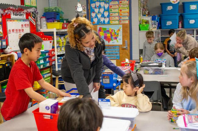 A UK student stands at a table in a second grade classroom and points to the students' work.