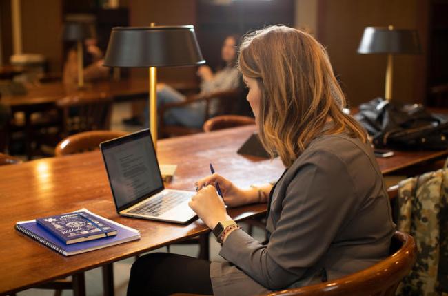 Photo of a woman sitting at a table in the William T. Young Library. She is typing on a laptop computer.