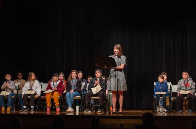 Photo of previous spelling bee. Students in the spelling bee are sitting in chairs on the stage behind a woman who is serving as emcee for the event, standing behind a podium and microphone.