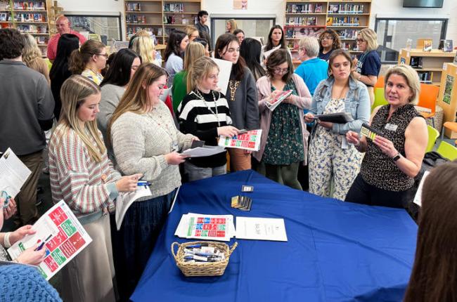 A group of students stands around a table with a blue table cloth. They are speaking to a hiring official from a nearby school district.