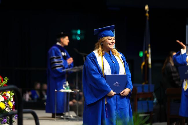 A woman stands on the University of Kentucky Commencement stage. She is wearing a cap and gown and smiling into the distance