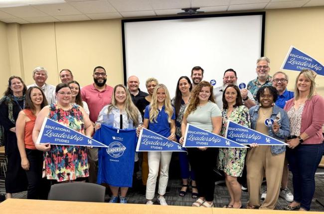 Large group of students and faculty stand in rows, holding up pennants and t-shirts that say UK College of Education