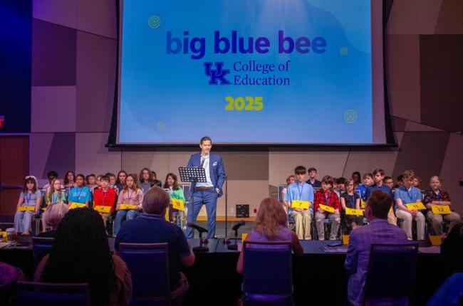 About 40 students sit in chairs in rows on a stage as an announcer introduces the 2025 spelling bee