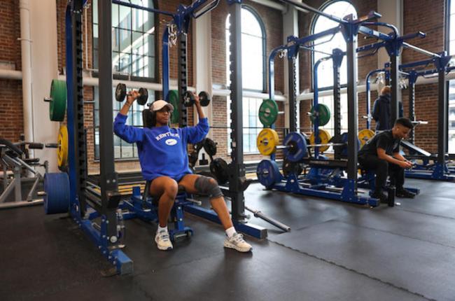 A woman sits on a bench and lift weights at Alumni Gym