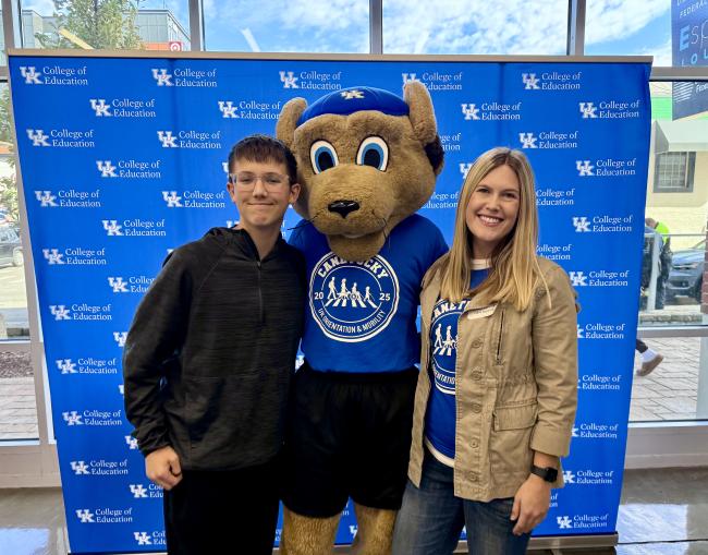 Two people participating in CaneTucky pose with the UK mascot Scratch in front of a UK College of Education blue backdrop