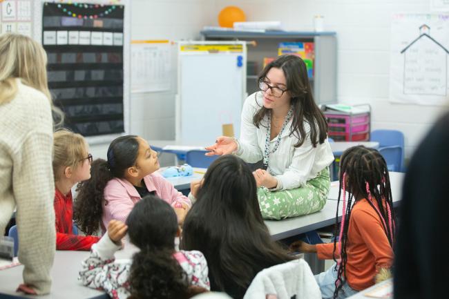 A UK College of Education student in a white shirt sits on a table, surrounded by elementary students listening to her talk