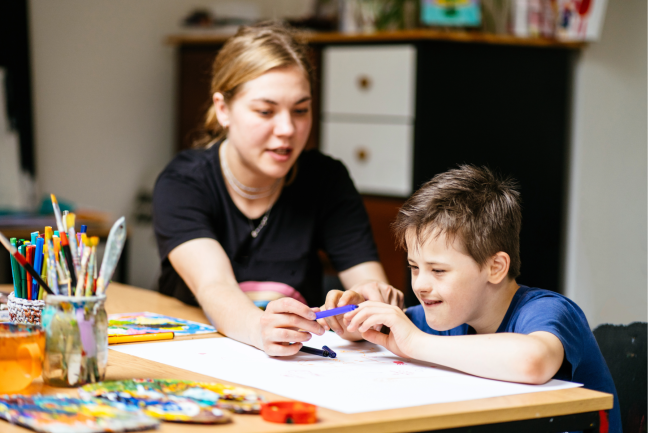 A woman sits next to a child at a school desk and assists the child with school work