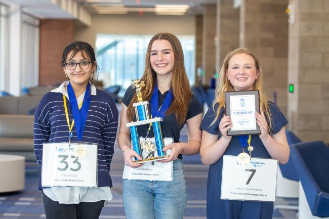 Three students pose for a photo. The girl in the middle holds a trophy as the spelling bee champion.