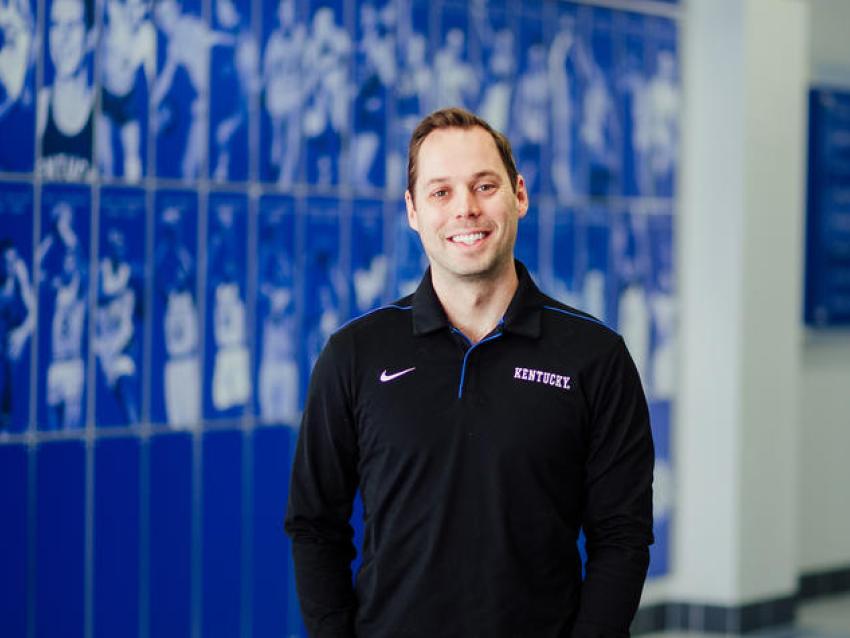 Man poses for a photo in front of a royal blue wall with images of student athletes on the wall