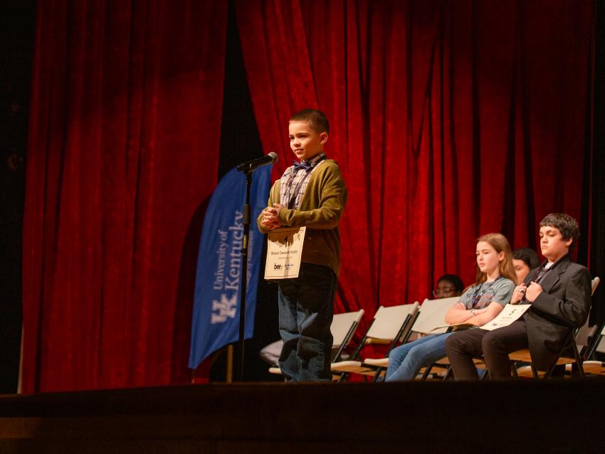 Boy stands behind microphone to spell a word in a 2024 spelling bee