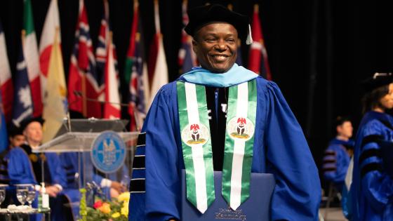 Photo of man wearing doctoral regalia. He is standing on stage at the University of Kentucky Commencement ceremony. There are flags on the stage behind him.