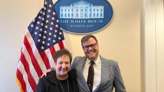 Two people standing in front of an American flag and a sign that says The White House