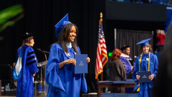 A woman in a blue cap and gown standing on a stage at commencement and holding up her diploma from the University of Kentucky.