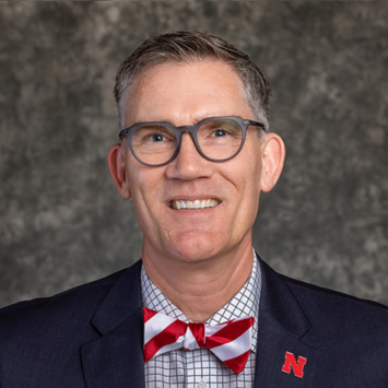 Photo of Nick Pace against grey background, wearing black jacket and red and white striped bow tie and glasses
