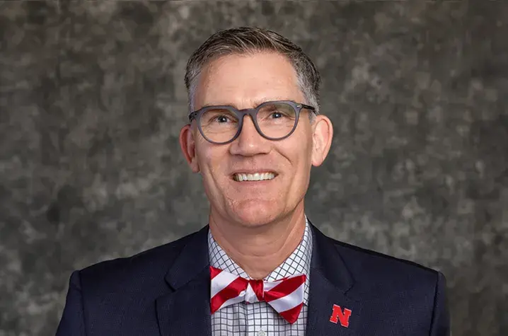 Photo of Nick Pace against grey background, wearing black jacket and red and white striped bow tie and glasses